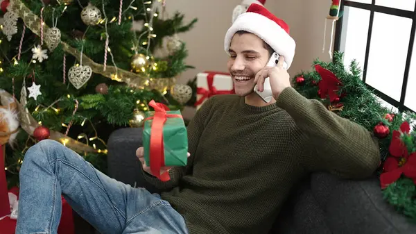 若いヒスパニックの男は 自宅でクリスマスプレゼントを持ってソファーに座ってスマートフォンで話します — ストック写真