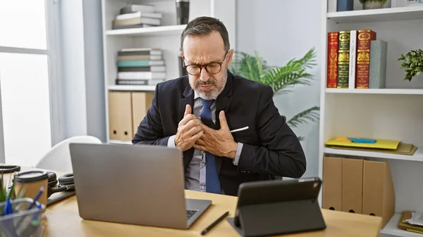 Stressed middle-aged man with grey hair, working hard in business, coughing at his office desk despite illness