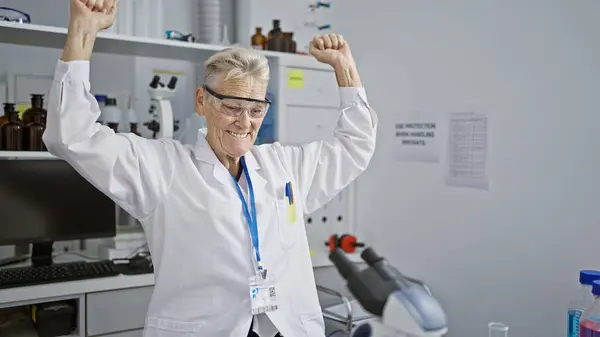 優勝を祝い 灰色の髪の女性科学者は研究室の顕微鏡で働いている間すべての笑顔で 研究医学の中心で熱意に満ちています — ストック写真