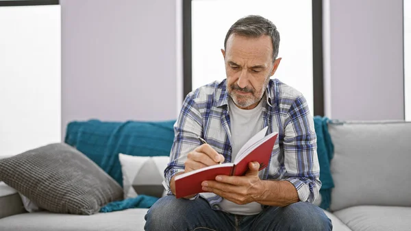 家庭で快適にリラックスして灰色の髪を持つ中年男性は ソファーで読書に魅了され 文学に深く集中しました — ストック写真