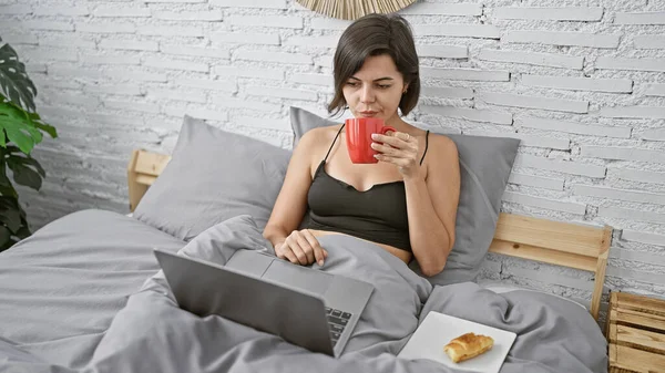 Beautiful young hispanic woman chilling in her bedroom, having a relaxed morning with breakfast in bed, using laptop and enjoying some online music.
