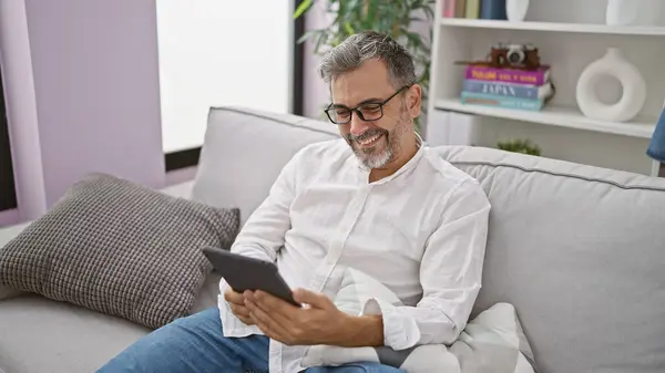 Confident young hispanic grey-haired man enjoys using touchpad while comfortably sitting on sofa and relaxing at home, exhibiting positive lifestyle indoors.