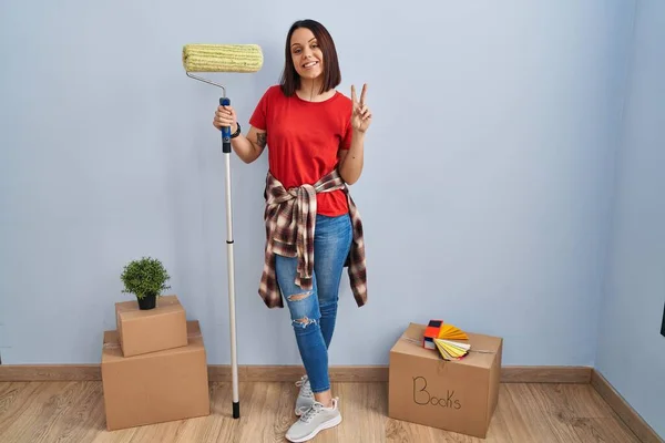 Young hispanic woman painting home walls with paint roller smiling with happy face winking at the camera doing victory sign with fingers. number two.