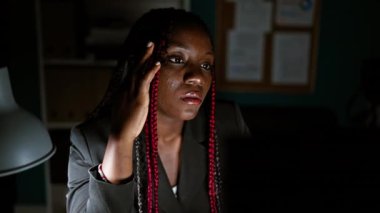 Yorgun afro-amerikan kadın işçi, iş kıyafetleri içinde, örgüler sıkı çekti, bilgisayarla aydınlatılmış çalışma odasında internet stresinde geziniyor, gece boyunca korkunç bir baş ağrısıyla mücadele ediyor.