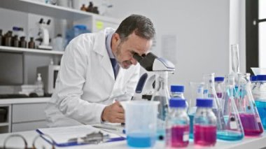 Beyaz saçlı orta yaşlı bir adam laboratuardaki panoda mikroskop kullanıyor.