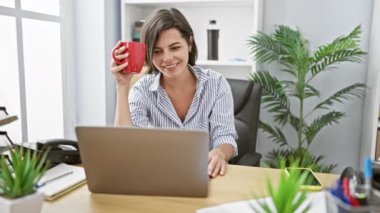 Güzel gülüşü ve kısa saçı olan, kendine güvenen genç İspanyol kadın patron, sabahın erken saatlerinde ofis masasında çalışırken bilgisayarını online iş başarısı için kullanırken kahvesini içiyor..
