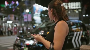 Güzel İspanyol kadın gece Tokyo sokaklarında taksi bekliyor, telefon uygulaması kullanıyor ve şehir ışıklarının tadını çıkarıyor.