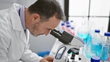 Gri saçlı, gülümseyen orta yaşlı bir adam laboratuarında mikroskop kullanarak bilimi neşeyle kucaklıyor.