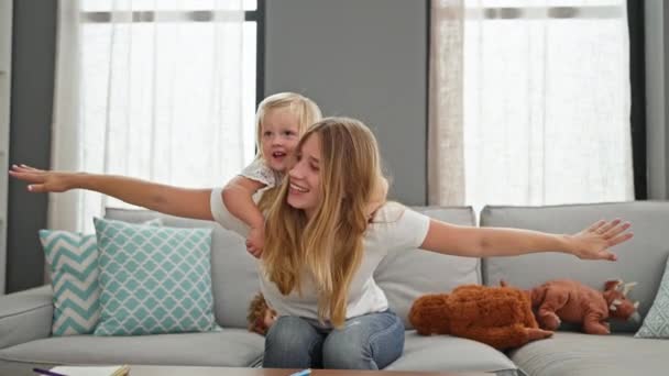 自宅で快適にソファーに座ってリラックスした慎重な母親 彼女の小さな娘を彼女の背中に遊ばせて 家族は幸せを放射します — ストック動画