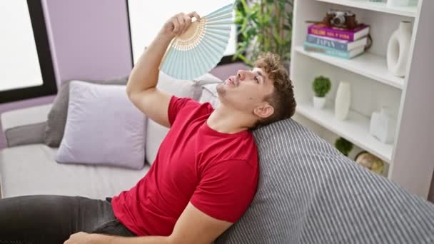 熱い天候の傷 若いヒスパニックの男は苦しみ 自宅でソファーに座ってハンドファンで自分自身をファニングし 屋内熱の苦労を露出 — ストック動画