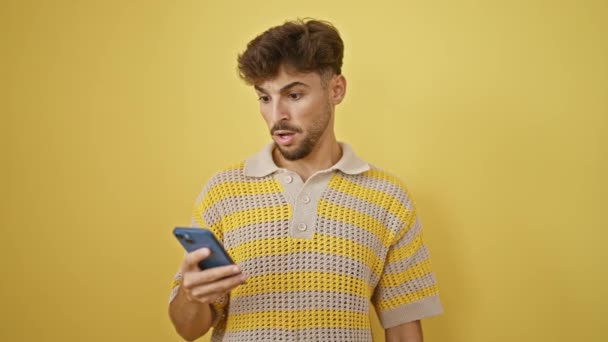 年轻的阿拉伯男子 留着胡子 睁大了眼睛 惊讶地站在那里 看着自己的智能手机 用孤立的黄色墙壁背景发短信 这是一个难以置信的信息吗 — 图库视频影像