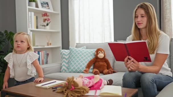 コーカサス人の母親と彼女の小さな少女は 快適で親しみやすい屋内設定の中で 自宅で暖かい物語の本に吸収され 喜んでノートに描かれています — ストック動画