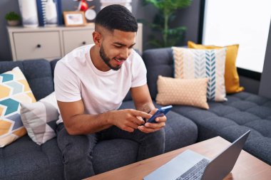 Evdeki kanepede oturan akıllı telefon kullanan genç latin adam.