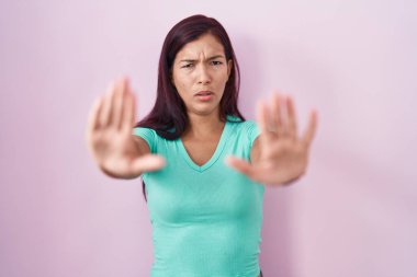 Young hispanic woman standing over pink background doing stop gesture with hands palms, angry and frustration expression 