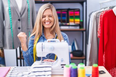 Dikiş makinesi kullanan sarışın kadın terzi çok mutlu ve heyecanlı bir şekilde kollarını kaldırıp, gülümseyerek ve başarı için bağırarak kazanan jestler yapıyor. kutlama konsepti. 