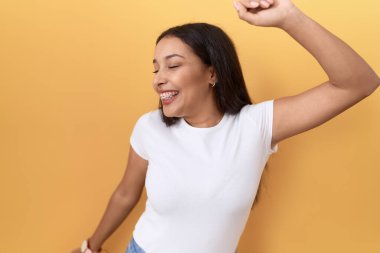 Sarı arka planda günlük beyaz tişört giyen genç Arap kadın mutlu ve neşeli dans ediyor, hareketli ve kendinden emin bir şekilde müzik dinliyor. 