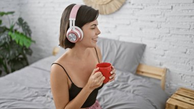Sabah saadeti, kendine güvenen, güzel, genç İspanyol kadın espresso yudumluyor, yatakta müzik dinliyor, rahat pijama ve battaniyeye sarılmış, gülümsüyor, evinde rahat bir şekilde dinleniyor.