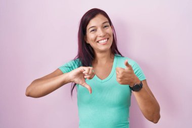 Latin kökenli genç bir kadın pembe arka planda dikiliyor. Baş parmaklarını yukarı aşağı hareket ettiriyor. Anlaşamamak ve anlaşamamak. çılgın çatışma 