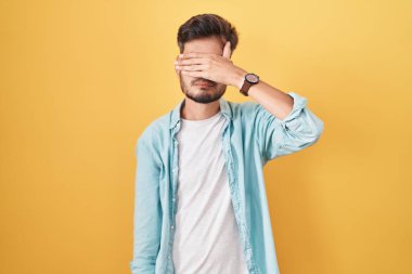 Sarı arka planda dikilen İspanyol dövmeli genç adam gözlerini eliyle kapatıyor, ciddi ve üzgün görünüyor. Görme, saklama ve reddetme konsepti 