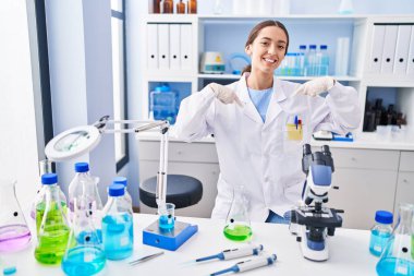 Young brunette woman working at scientist laboratory looking confident with smile on face, pointing oneself with fingers proud and happy. 