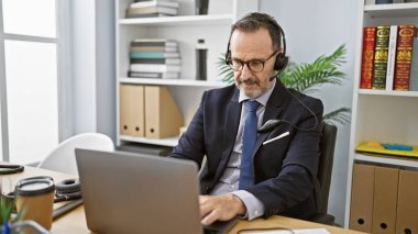 Beyaz saçlı, ispanyol orta yaşlı bir adam, ofisteki işine odaklanmış, profesyonel olarak bir müşteriyle konuşuyor, dizüstü bilgisayarla yardım sağlıyor. Kulaklık kullanarak konsantre ve başarılı bir işçi.