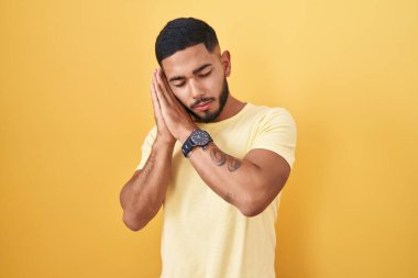 Sarı arka planda dikilen İspanyol genç adam yorgun uyuyor. Hayal kuruyor ve elleri ile birlikte poz veriyor. Gözleri kapalı gülümsüyor.. 