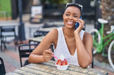 Afro-Amerikalı kadın dondurma yiyor, kafenin terasında akıllı telefondan konuşuyor.