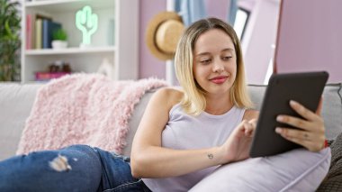Young blonde woman using touchpad sitting on sofa at home