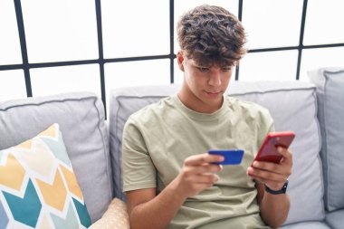 Genç İspanyol genç akıllı telefon ve kredi kartı kullanıyor. Evdeki kanepede oturuyor.