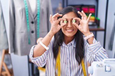 Young beautiful hispanic woman tailor smiling confident holding buttons over eyes at clothing factory