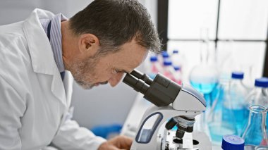 Gri saçlı yaşlı bir adam, laboratuarında çalışan bir bilim adamı, tıbbi keşif için mikroskoba bakıyor.