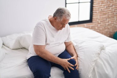 Middle age grey-haired man suffering for knee injury sitting on bed at bedroom