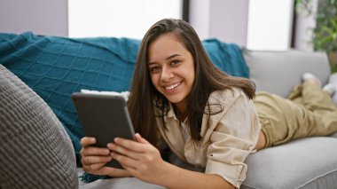 Evdeki kanepede uzanmış dokunmatik ped kullanan genç, güzel İspanyol kadın.