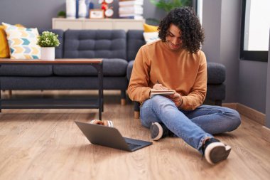 Young latin man using laptop writing on notebook at home