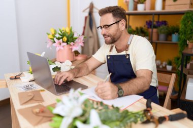 Middle age man florist using laptop writing on notebook at flower shop