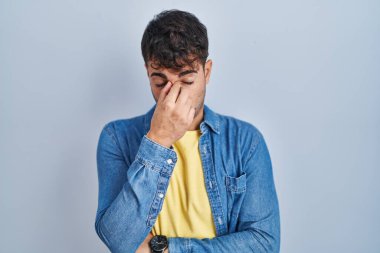 Young hispanic man standing over blue background tired rubbing nose and eyes feeling fatigue and headache. stress and frustration concept. 