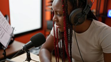 Kendine güvenen Afro-Amerikan kadın muhabir. Kapalı bir müzik stüdyosunda canlı yayında konuşuyor. Radyodan, kulaklıklarla haberleri yayınlıyor.
