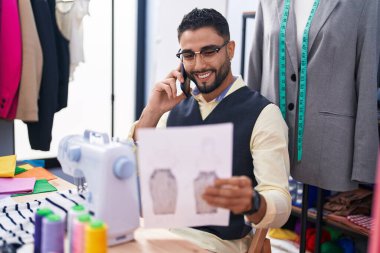 Genç Arap terzi akıllı telefondan konuşuyor giyim fabrikasında kıyafet tasarımı yapıyor.