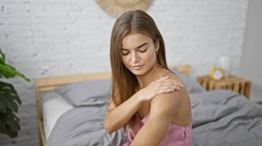 Güzel, genç, İspanyol bir kadın, rahat bir şekilde yatağında oturuyor, rahat bir gülümseme ile cilt bakımını uyguluyor..