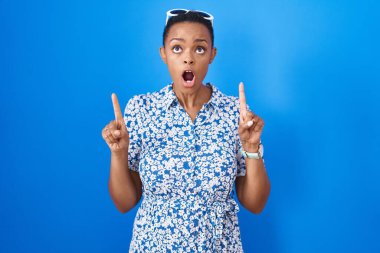 Mavi arka plan üzerinde duran Afrikalı Amerikalı kadın şaşırmış ve şaşırmış bir şekilde yukarı bakıyor ve parmaklarıyla işaret ediyor ve kollarını kaldırıyor.. 