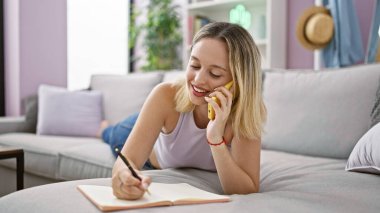 Not defterine yazan genç sarışın kadın evde akıllı telefondan konuşuyor.