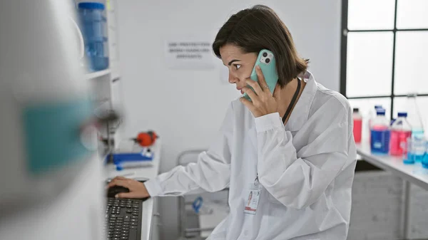 Young, beautiful hispanic woman scientist earnestly immersed in a tech-filled conversation on smartphone while working on computer in bustling lab