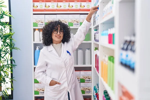 Young middle eastern woman pharmacist smiling confident holding bottle of shelving at pharmacy