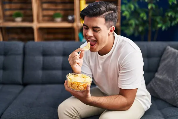 Young hispanic man eating chips potatoes sitting on sofa at home