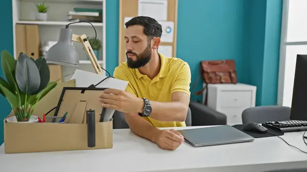 Young hispanic man being fired sitting on desk looking belongings package at the office