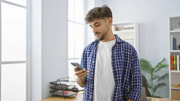 Hardworking young arab man immersed in his professional world, concentrated on typing message on smartphone at the office, epitomizing a successful business worker lifestyle.