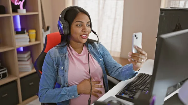 Heartwarming moment of a young, pregnant gaming streamer, playfully touching her belly and smiling during a night video call in her gaming room, before launching her new game.