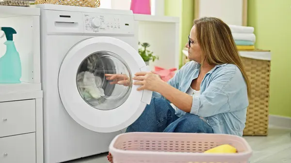 Middle age hispanic woman washing clothes sitting on floor at laundry room