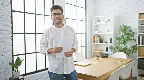 一个成功的年轻人在工作时 信心十足地拿着商业文件 在办公室的背景下微笑 由此散发出欢乐的光芒 — 图库照片