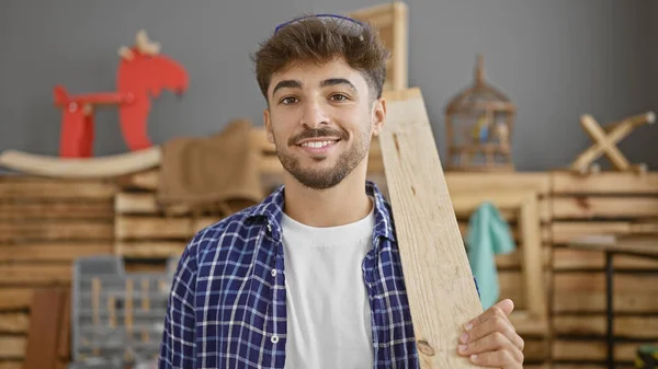 Smiling young arab man at work, professional carpenter confidently handles wood plank in his carpentry studio
